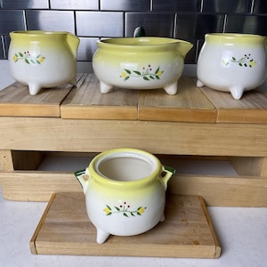 May include: A collection of vintage ceramic pots with a white base and yellow rim. Each pot features a floral design and rests on small, white feet. The pots are arranged on a wooden display, showcasing their unique shapes and sizes.