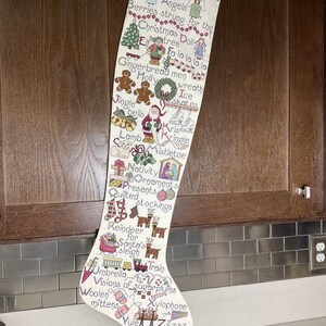 May include: A cream-colored Christmas stocking with cross-stitched holiday imagery and the year "1990." The design includes angels, gingerbread men, reindeer, and other festive elements. The stocking is hanging against a wooden cabinet.