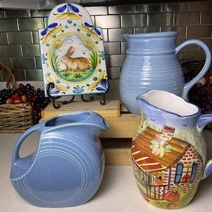 May include: An assortment of ceramic pitchers and a decorative tile. The pitchers are in shades of blue, with one featuring a house design. The tile depicts a rabbit in a garden setting, with a floral border. A rustic kitchen setting.