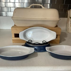 May include: A set of kitchenware including a large, rectangular, beige stoneware bread baker with a lid and three oval-shaped baking dishes. The dishes feature a glossy white interior and a deep blue exterior with integrated side handles.