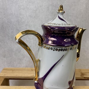 May include: A white porcelain teapot with a purple and gold design. The teapot has a curved handle and spout, and a lid with a gold finial. The purple design swirls down the side of the teapot.