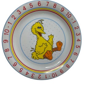 May include: A round white ceramic plate featuring a central illustration of a yellow bird character with orange feet. The rim is decorated with a repeating sequence of red numbers from one to ten, separated by dots, and bordered by yellow and red rings.