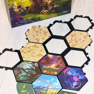 May include: A hexagonal game board with black borders and colourful illustrations of a forest, a castle, a shop, and a cave. The board is made up of six hexagonal tiles and a circular outer ring with symbols and numbers.