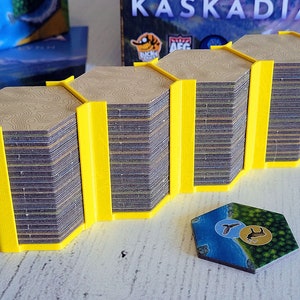 Könnte beinhalten: Ein gelber 3D-gedruckter Organizer hält Stapel sechseckiger Spielsteine. Die Spielsteine haben ein braunes topografisches Kartendesign. Ein einzelner Spielstein mit einer blau-grünen Landschaft und Tiersymbolen befindet sich im Vordergrund. Die Spielbox ist im Hintergrund.