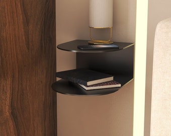 May include: A black floating shelf with two tiers, mounted on a wall. The shelf is holding a lamp with a white shade and a stack of books. The shelf is in a bedroom setting.