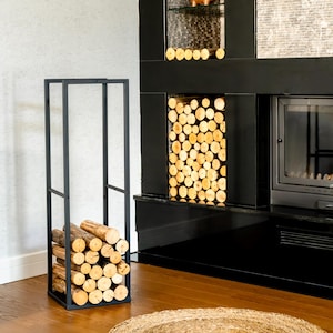 Könnte beinhalten: Ein schwarzer Metall-Holzhalter, gefüllt mit geschnittenem Brennholz, steht auf einem Holzboden. Der Halter ist rechteckig und minimalistisch gestaltet. Im Hintergrund befindet sich ein schwarzer Kamin mit Glastür und dekorativer Nische.