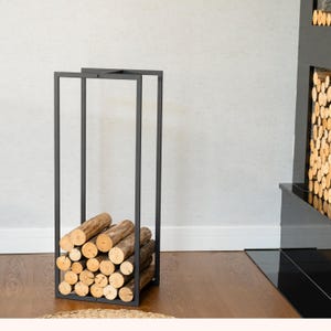 Könnte beinhalten: Ein schwarzer Metall-Holzhalter, gefüllt mit geschnittenen Holzscheiten. Der rechteckige Halter hat ein minimalistisches Design mit einem stabilen Rahmen. Die Holzscheite sind ordentlich gestapelt, wobei die natürliche Holzmaserung und die kreisförmigen Endmuster zur Geltung kommen. Der Halter steht auf einem Holzboden.