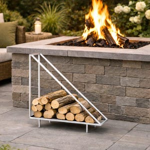 Könnte beinhalten: Ein weißer Metall-Holzständer hält einen Stapel Holzscheite, positioniert vor einer Feuerstelle aus Stein mit einem brennenden Feuer. Die Feuerstelle befindet sich in einem Terrassenbereich mit Gartenmöbeln und Lichterketten.
