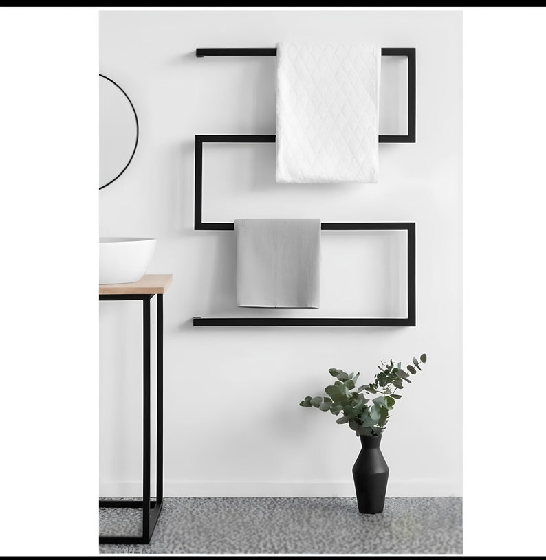 May include: A contemporary black metal towel rail with two towels. One towel is white with a quilted design, the other is grey. The rail is on a white wall, with a black vase and greenery in the foreground. A minimalist bathroom design.
