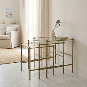 May include: A trio of nesting tables featuring gold-toned metal frames and clear glass surfaces. The tables are arranged in a nested configuration, with the largest table at the rear and the smallest at the front. The tables are in a living room setting.