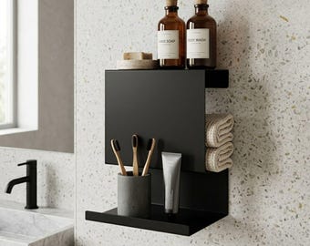 Puede incluir: Estantería de baño de metal negro con dos botellas de vidrio marrón etiquetadas como "Hand Soap" y "Body Wash". La estantería contiene un soporte gris para cepillos de dientes con tres cepillos, un tubo de pasta de dientes y toallas beige dobladas.