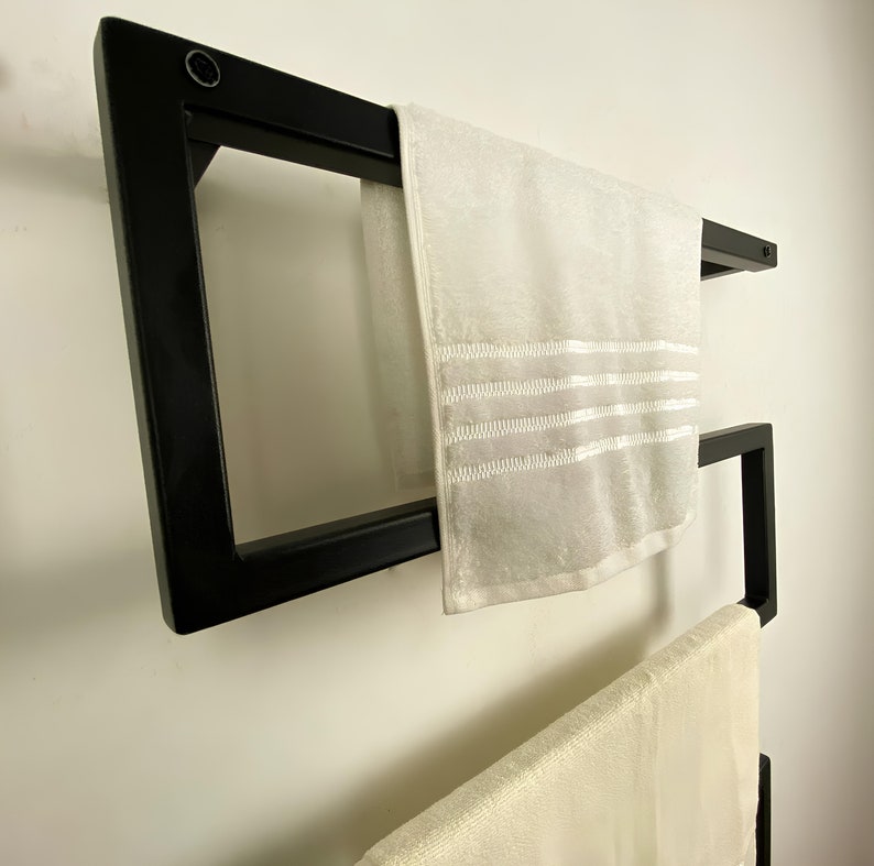 May include: A contemporary black towel rail fixed to a white wall, displaying two white towels. The rail features a geometric design with square and angled sections. The towels have decorative stripes.