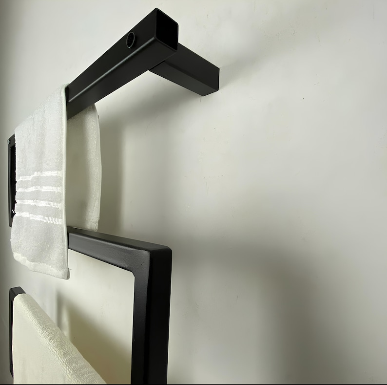 May include: A contemporary black metal towel rail with a geometric design. The rail holds two white towels with striped detailing. The rail is mounted on a white wall, creating a minimalist aesthetic.