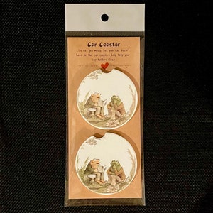 May include: Two round car coasters with a frog theme. The coasters feature two frogs sitting on a log, one reading a book and the other listening. The coasters are white with a brown border and a green and brown frog design. The packaging is clear plastic with a brown card backing. The card has the text "Car Coaster" and "Life can get messy, but your car doesn't have to. Our car coasters help keep your cup holders clean."