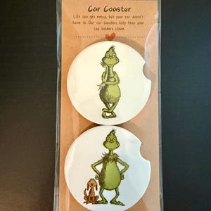 May include: Two white car coasters with green Grinch illustrations. One Grinch is standing, arms akimbo, with a dog. The other Grinch is standing with arms crossed. The packaging reads "Car Coaster".
