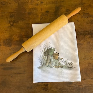 May include: A wooden rolling pin rests on a white waffle-weave kitchen towel. The towel features a whimsical illustration of two frogs and a snail. The rolling pin is light brown and appears to be made of a single piece of wood.