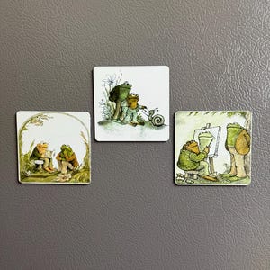 May include: Three square magnets featuring illustrations of frogs. One shows two frogs sitting, another depicts frogs on a scooter, and the third shows a frog painting. The artwork has a whimsical, storybook style with green and brown tones.