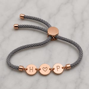 May include: A grey cord bracelet featuring three rose gold circular charms. The outer charms are engraved with the letters H and P, while the center charm displays a heart symbol. The bracelet includes a rose gold adjustable sliding bead closure.