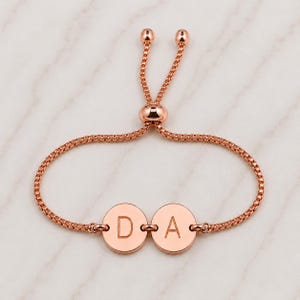 May include: A rose gold-toned adjustable bracelet featuring a box chain and two circular charms. Each charm is engraved with a single uppercase letter, specifically the letters D and A, positioned side by side in the center of the bracelet.