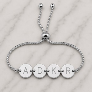 May include: A silver-toned adjustable bracelet featuring a box chain and four circular charms. Each charm is engraved with a single uppercase letter, spelling out A, D, K, and R. The bracelet includes a sliding bead mechanism with two small spheres at the ends of the chain for size adjustment.