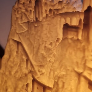 May include: A 3D printed lithophane lamp featuring a detailed relief of a mountain landscape with architectural structures, including towers and arched bridges, illuminated from within to create a warm, glowing amber light effect.