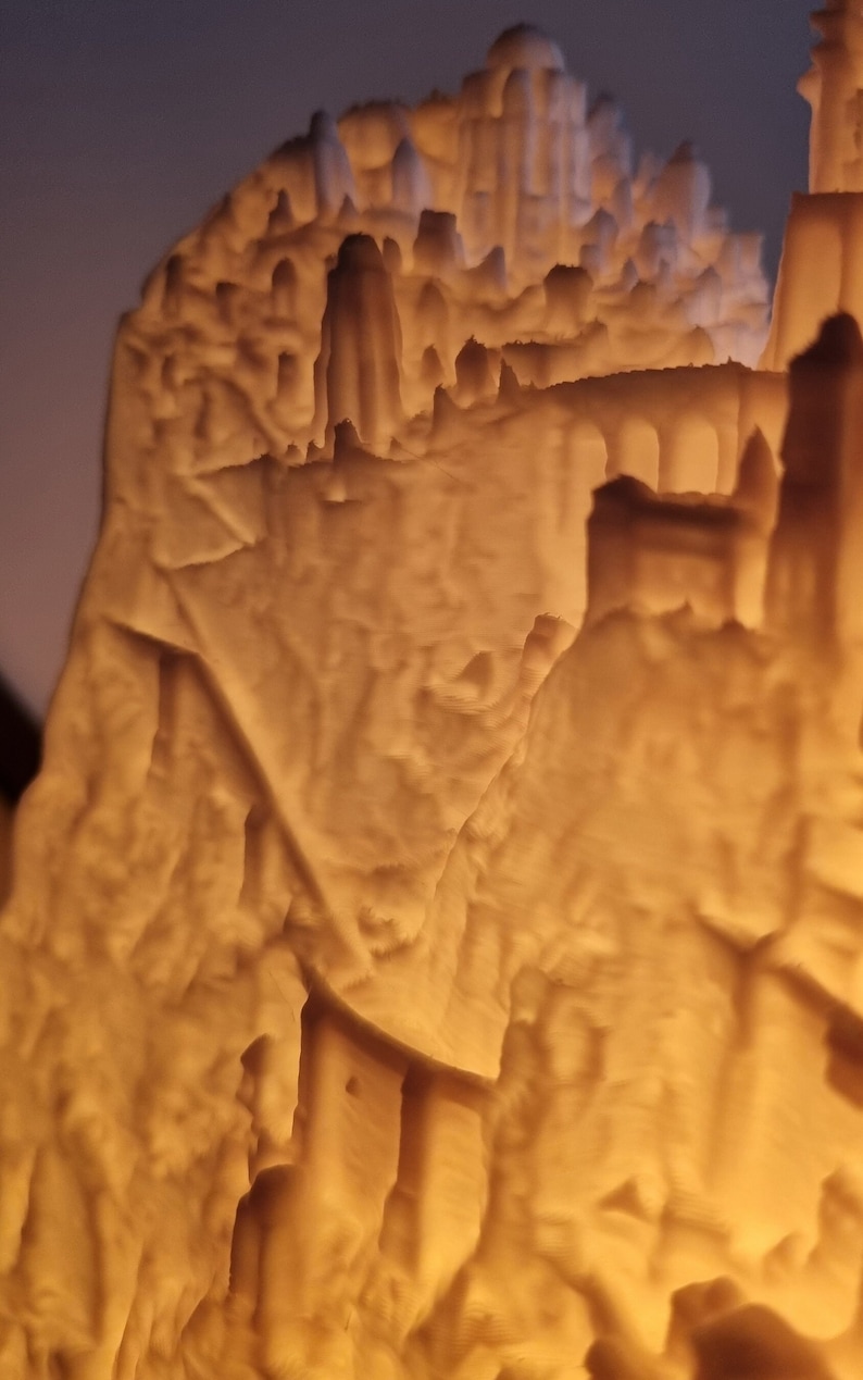 May include: A 3D printed lithophane lamp featuring a detailed relief of a mountain landscape with architectural structures, including towers and arched bridges, illuminated from within to create a warm, glowing amber light effect.
