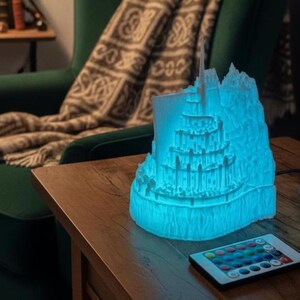 May include: A white 3D printed lamp shaped like a multi-tiered castle structure, illuminated from within by a bright blue light. The lamp sits on a wooden surface next to a small rectangular remote control with multiple colored buttons for light settings.