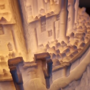 May include: A 3D printed off-white lamp featuring a detailed architectural structure with tall columns, intricate wall textures, and a crenelated perimeter wall. The object is illuminated from within, casting a warm glow through the translucent material.
