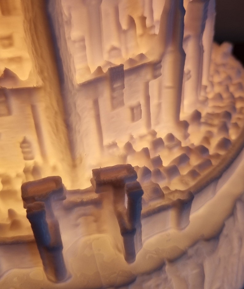 May include: A 3D printed off-white lamp featuring a detailed architectural structure with tall columns, intricate wall textures, and a crenelated perimeter wall. The object is illuminated from within, casting a warm glow through the translucent material.