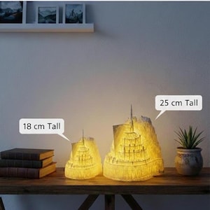 May include: Two illuminated, off-white, castle-shaped lamps with intricate architectural details. The smaller lamp is 18 cm tall, and the larger lamp is 25 cm tall. Both emit a warm, soft yellow glow from within, highlighting the textured structure.