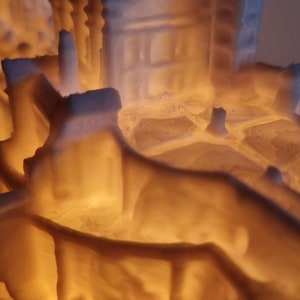 May include: A 3D printed white architectural model of a building complex illuminated from within by a warm orange light. The structure features intricate walls, towers, and layered surfaces that cast soft shadows, highlighting the textured finish.