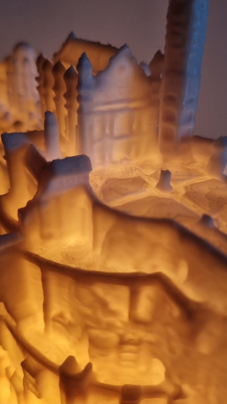 May include: A 3D printed white architectural model of a building complex illuminated from within by a warm orange light. The structure features intricate walls, towers, and layered surfaces that cast soft shadows, highlighting the textured finish.