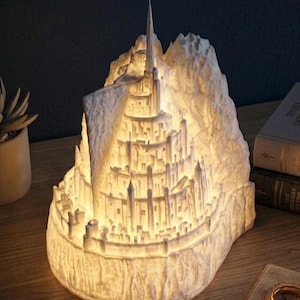 May include: A white, illuminated 3D-printed sculpture depicting a multi-tiered fortress city built into a steep mountain. The structure features numerous towers, walls, and levels, glowing from within to highlight the intricate architectural details.