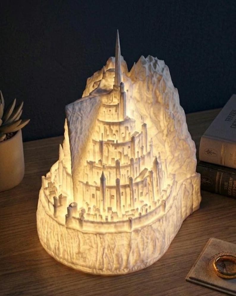 May include: A white, illuminated 3D-printed sculpture depicting a multi-tiered fortress city built into a steep mountain. The structure features numerous towers, walls, and levels, glowing from within to highlight the intricate architectural details.