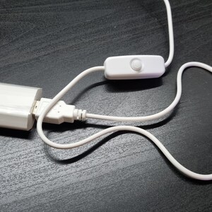May include: A white power adapter with a two-prong plug and a USB port, connected to a white cable featuring an inline rocker switch. The cable is flexible and white, extending from the USB port to the switch and continuing beyond the switch.