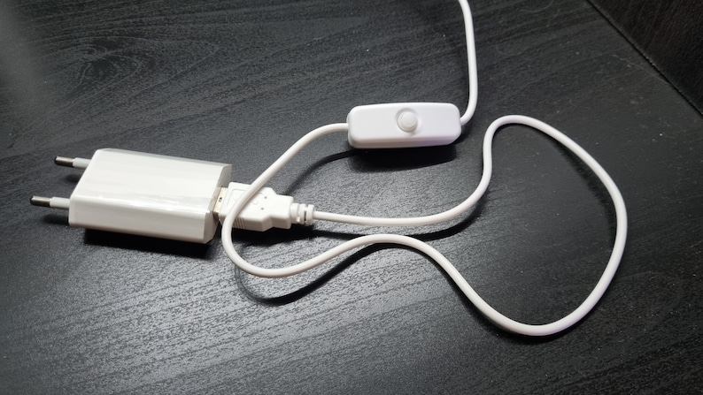 May include: A white power adapter with a two-prong plug and a USB port, connected to a white cable featuring an inline rocker switch. The cable is flexible and white, extending from the USB port to the switch and continuing beyond the switch.