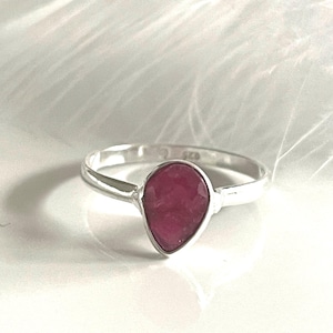 Könnte beinhalten: Ein silberner Ring mit einem tropfenförmigen, tiefroten Edelstein. Der Ring hat ein dünnes, poliertes Band, und der Edelstein ist in einer einfachen Lünette gefasst. Der Ring wird auf einer weißen Oberfläche präsentiert.