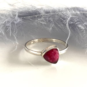 Könnte beinhalten: Ein silberner Ring mit einem rosa dreieckigen Edelstein. Der Ring liegt auf einem weißen Hintergrund mit einer Feder.