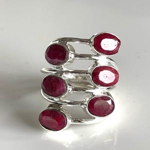 Könnte beinhalten: Ein silberner Ring mit sechs ovalen, tiefroten Rubinen. Die Rubine sind in einem spiralförmigen Design angeordnet, wobei jeder Stein von einer einfachen Fassung gehalten wird. Der Ring hat eine polierte Oberfläche und wird auf weißem Untergrund präsentiert.