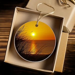 May include: Round ceramic ornament featuring a sunset over the ocean scene. The ornament depicts a bright orange and yellow sunset with a silhouette of grass in the foreground.