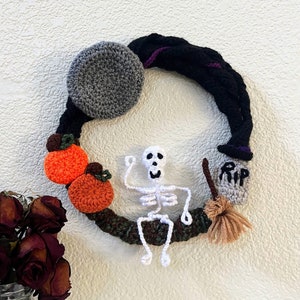 May include: A handmade Halloween wreath featuring a black and purple braided design. The wreath includes a gray moon, orange pumpkins, a white skeleton, a tombstone with "RIP" text, and a broom. The wreath is a festive decoration.