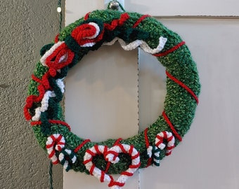 May include: A festive, handmade Christmas wreath. The green crocheted base is decorated with red and white details, including candy canes and heart shapes. The wreath hangs on a white door, with fairy lights above.
