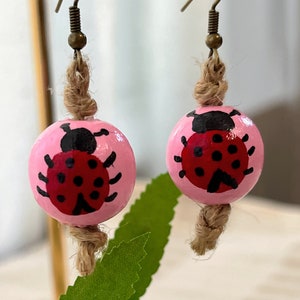 May include: A pair of pink wooden earrings with a ladybug painted on each. The earrings are attached to brown twine and have a gold hook.