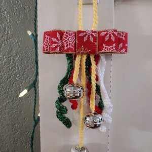 May include: A Christmas ornament featuring a red ribbon with white snowflakes, gold trim, and a yellow crocheted loop. Silver bells hang below with green, red, and white crocheted accents. The ornament is hanging on a white door.