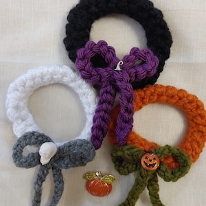 May include: A collection of handmade Halloween-themed crochet wreaths in black, white, and orange. Each wreath features a decorative bow in coordinating colors, with embellishments like a skull, a silver bell, and a pumpkin. A small pumpkin pin is also included.