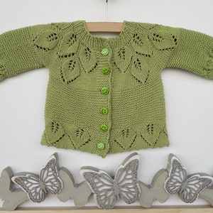 May include: A green knitted baby cardigan with a leaf pattern and green buttons. The cardigan is laid flat on a white surface with decorative wooden butterflies.