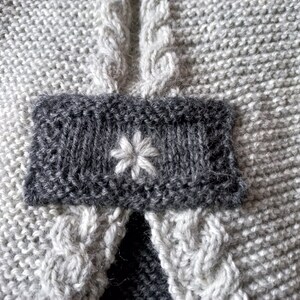 May include: A close-up of a gray knitted fabric with a dark gray patch featuring a white embroidered flower.