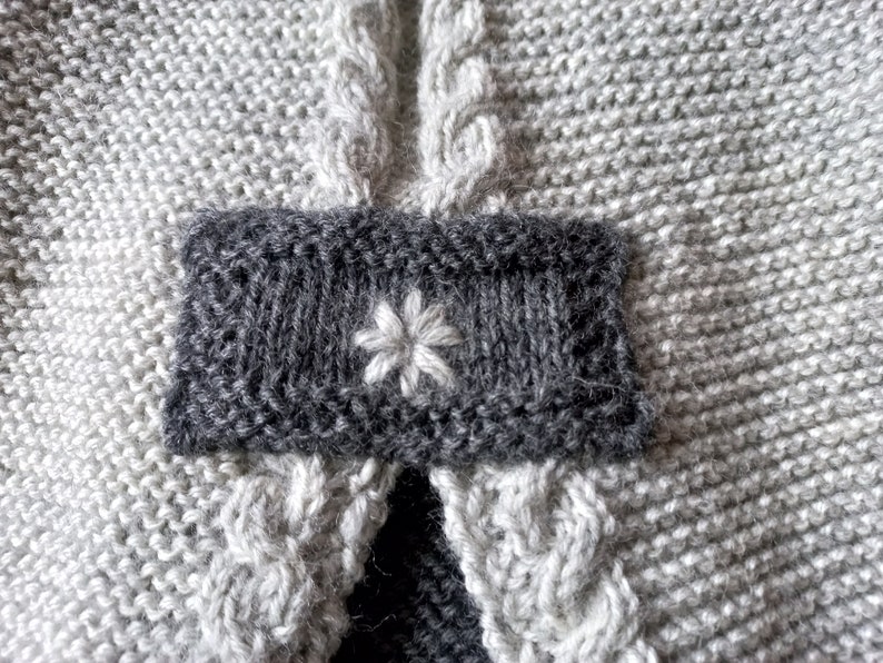 May include: A close-up of a gray knitted fabric with a dark gray patch featuring a white embroidered flower.