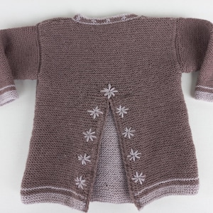 May include: A brown knitted cardigan with a split back, decorated with embroidered white flower accents. The cuffs and hem have a light purple trim. This handmade sweater is designed for a baby or toddler.