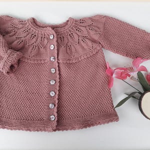 May include: A dusty rose-colored knitted cardigan with a round neckline and long sleeves. The yoke features a decorative leaf pattern, and the front has six clear buttons. The cardigan is a baby or toddler garment.